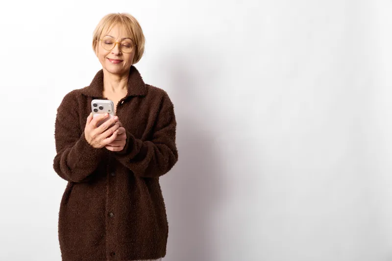 Elderly woman portrait indoors, smiling while holding smartphone, wearing brown coat, relaxed stance, clean white background, soft studio light, calm mood, gently