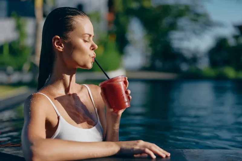 Young woman in white swimsuit relaxing in pool edge drinking red smoothie through straw sunny day outdoor lifestyle healthy refreshment