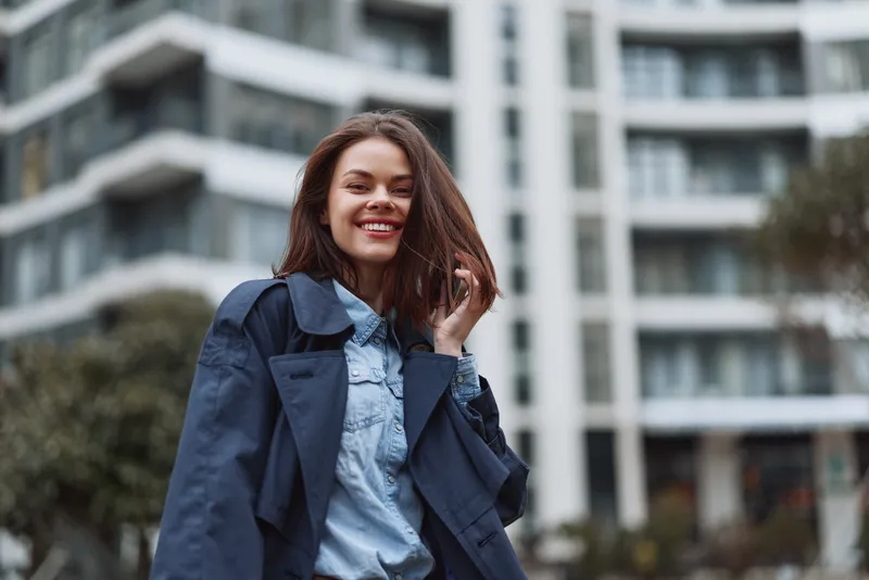 A smiling young woman in a denim shirt and navy jacket stands outdoors in a modern city, conveying confident casual style and urban positivity today.