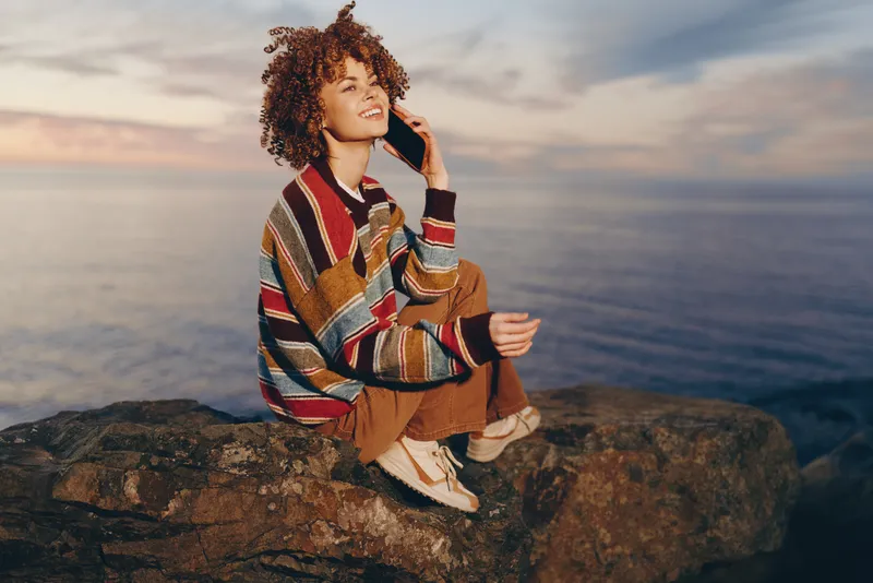 Woman with curly hair talking on phone outdoors near ocean at sunset, standing on rocky cliff, relaxed lifestyle moment, warm golden light, casual wear, travel mood, smiling