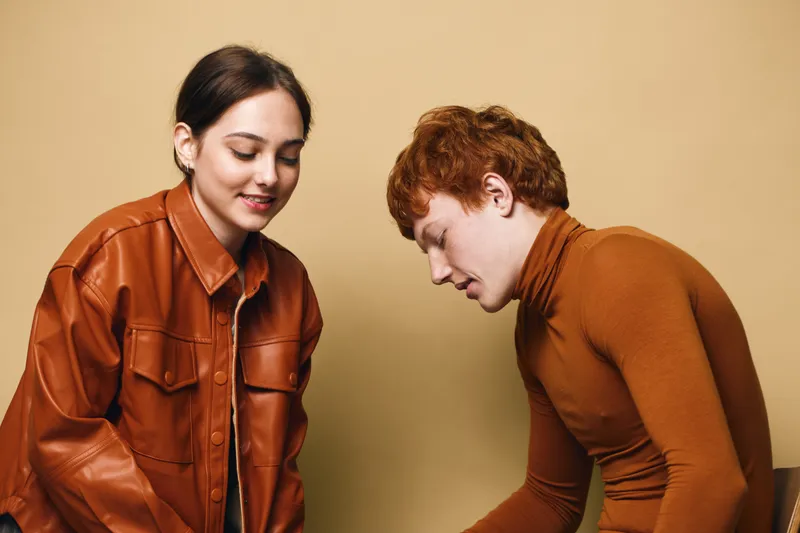 Young man and woman dressed in brown and orange clothing standing and looking downward with smiles on beige background for fashion style and casual portrait.