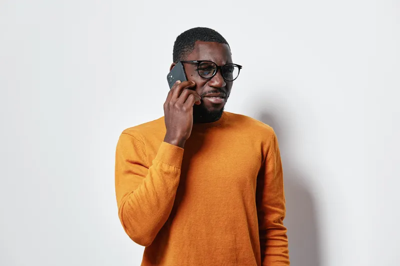 African man with dark skin wearing glasses and orange sweater talking on smartphone against white background. Casual communication, technology and portrait with confident expression.
