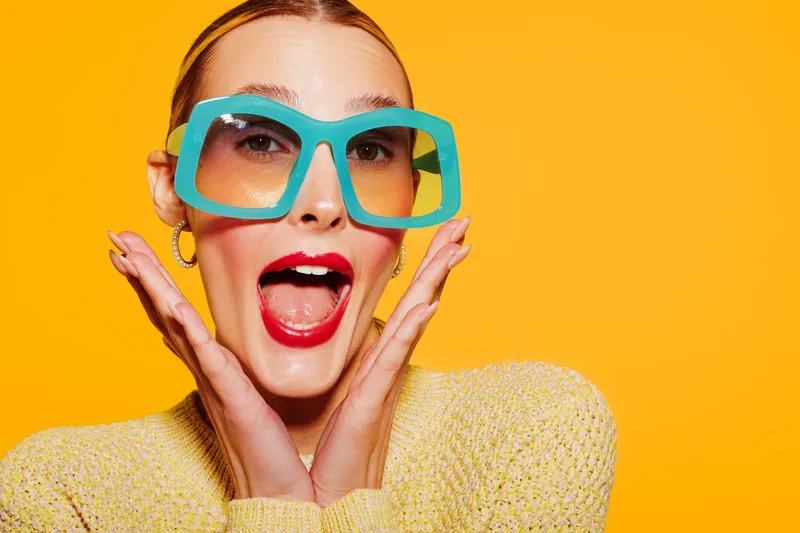 Excited woman with bright red lipstick and large turquoise glasses poses with open mouth and hands near face on vibrant yellow background. Concept of joy, fashion, and expression.