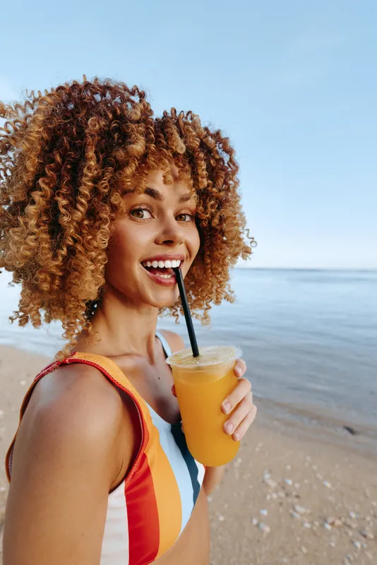 Young woman with curly hair enjoys a refreshing orange drink by the calm sea on a sunny day, smiling happily in a colorful striped swimsuit at the beach.