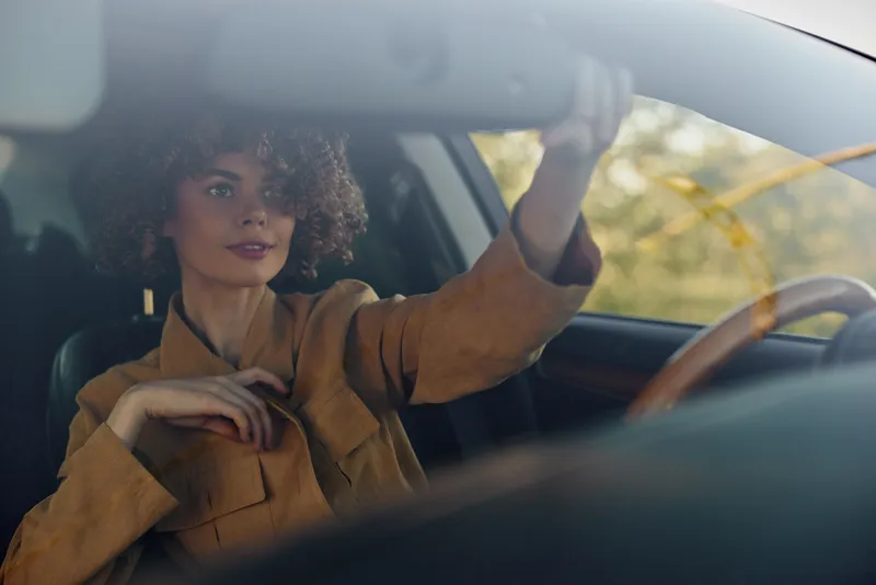 Cheerful woman driving a car, seated in warm autumn jacket, sunlight through window, confident pose, relaxed smile, soft breeze hinted, natural colors, urban road setting