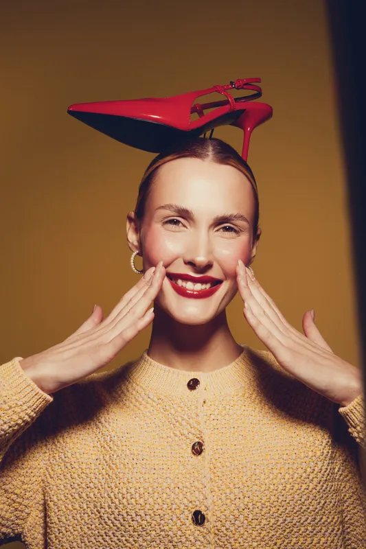 Smiling woman with red lipstick and hoop earrings balances red high heel shoe on head. She wears beige sweater and poses against yellow background. Concept of fashion and fun.