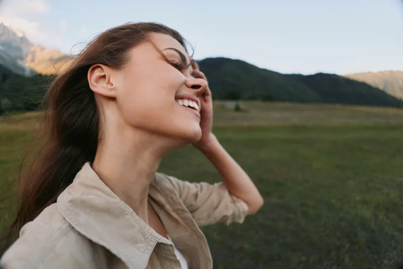 Portrait of a smiling woman enjoying nature in an open field, mountains in the background, conveying carefree outdoor lifestyle, personal happiness and natural serenity, peaceful moment.