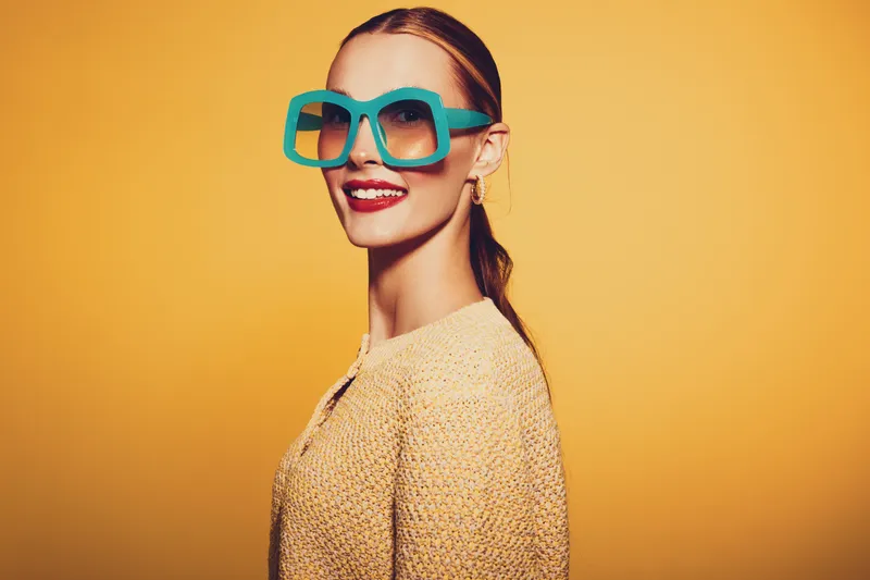 Smiling woman with large turquoise sunglasses and red lipstick poses against yellow background. Fashion portrait with bright colors and confident expression in studio lighting.