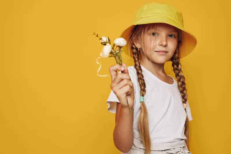 Young girl with braided hair wears a yellow hat and white t-shirt holding cotton flowers. Studio portrait on yellow background. Concept for childhood, innocence, nature.