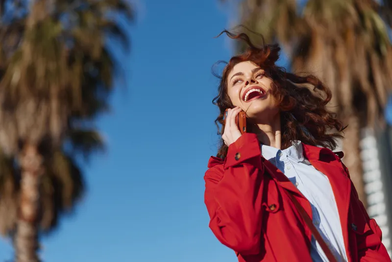 Smiling woman wearing a red jacket chats on her phone outdoors under a bright blue sky a candid moment of confidence and style in a casual setting.