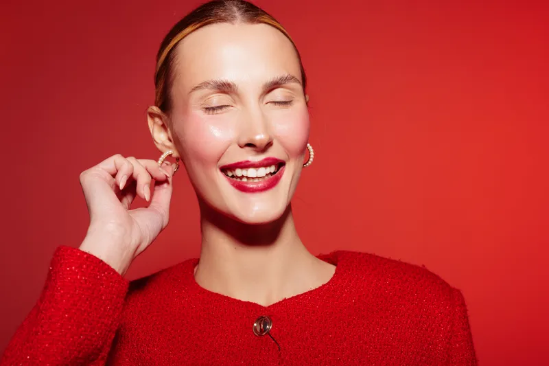Smiling woman with closed eyes wearing red sweater and gold earrings poses against red background. Concept of happiness, fashion, beauty, and joyful emotions in studio lighting.