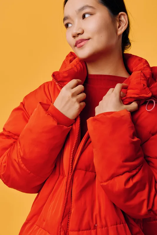Young woman wearing red puffer jacket looks to the side with a confident smile against yellow background. Casual style, winter clothing and lifestyle portrait with bright colors.
