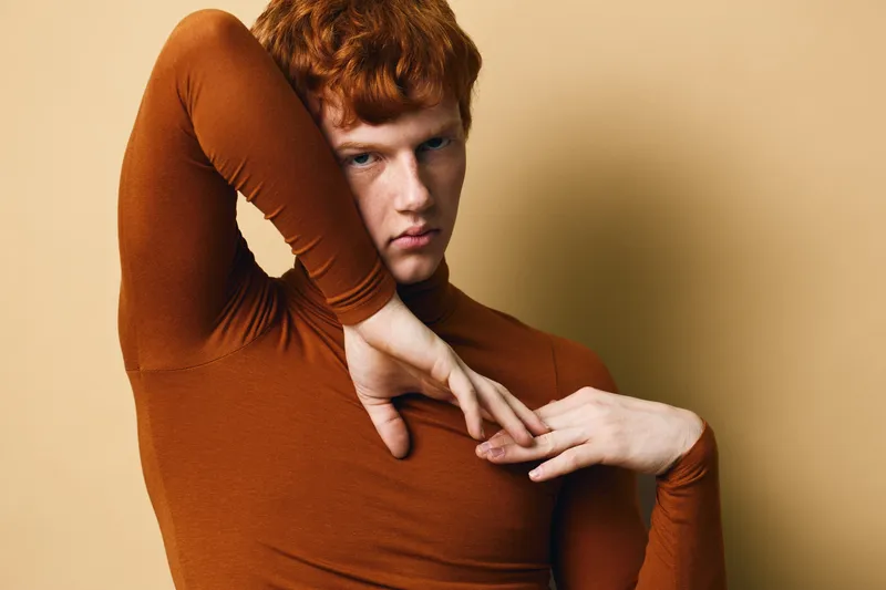 Redhead man posing with expressive hands and intense gaze wearing brown long sleeve shirt against beige background. Studio portrait with minimal style and dramatic mood.