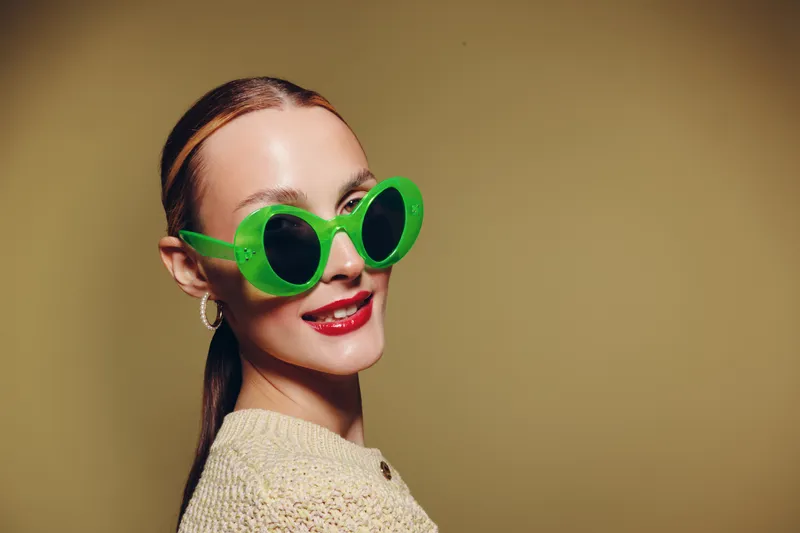 Young woman with green round sunglasses smiles confidently wearing beige knitted sweater on beige background. Concept of fashion, style, accessories, and modern portrait photography.