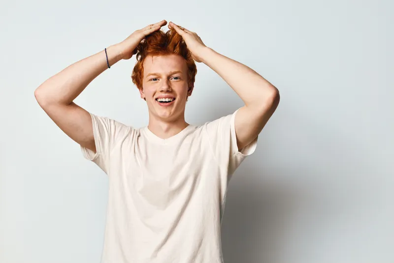Young man with red hair smiling and holding head in white t shirt against plain background. Emotion, casual style, happy expression, isolated studio portrait.