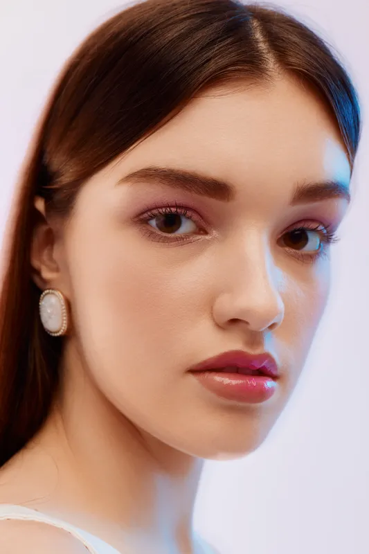 Close-up portrait of a young woman with smooth skin and glossy lips wearing elegant earrings. Soft lighting highlights her beauty and fashion style on a neutral background.