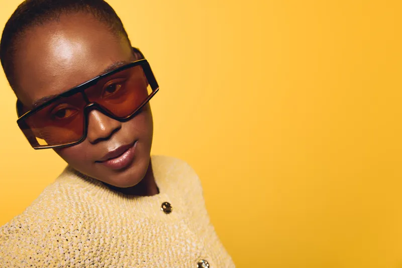 Stylish young black woman wearing oversized sunglasses and a beige sweater poses confidently against a vibrant yellow background. Concept of fashion, beauty, and modern style.