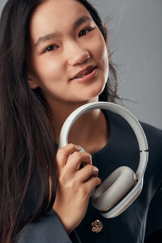 Young woman with long dark hair holding wireless headphones in hand looking at camera with soft smile against grey studio background. Casual clothing and modern audio device closeup portrait.