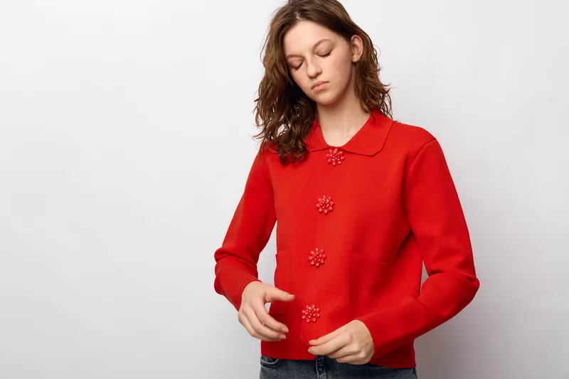 Young woman in red knitted sweater with floral buttons looks down thoughtfully on white background. Casual fashion, autumn style, and cozy clothing concept with natural light.