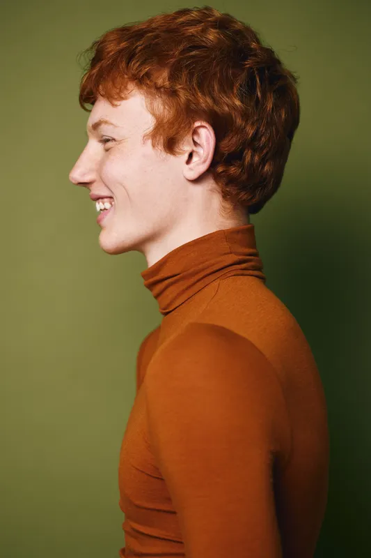 Young man with red hair and freckles wearing brown turtleneck sweater in profile smiling cheerfully against green background studio portrait of happy male model.