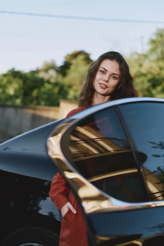 A young brunette woman stands beside a sleek car in a bright outdoor setting, exuding confidence and casual urban style