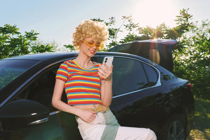 Smiling woman with curly blonde hair wearing a striped shirt stands beside a dark car, checking her smartphone. Retro color grade and bright natural light convey a stylish, carefree lifestyle mood.