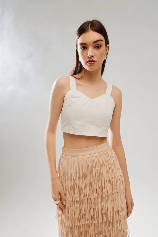 Young woman in a white crop top and beige fringed skirt poses in a studio with soft lighting. Concept of woman beauty fashion, elegance, and modern style.