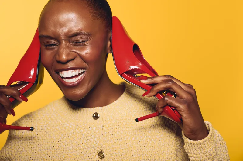 Smiling dark skinned African woman holding red high heels near ears on yellow background. Concept of fashion, joy, beauty, and playful emotions in studio portrait of black female model.