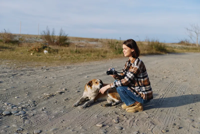 Woman crouching beside a dog on a sandy rural landscape, wearing a plaid shirt and jeans, holding a camera and capturing a candid moment of outdoor companionship.