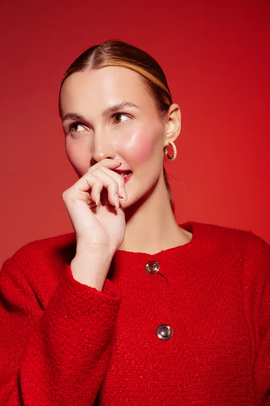 Young woman in red sweater poses thoughtfully against red background, with natural makeup and gold hoop earrings. Concept of fashion, style, and confident female portrait.