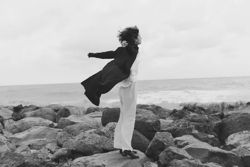 Sea breeze, woman on jagged rocks, coat billowing, capturing freedom and balance against a windy shore in monochrome landscape
