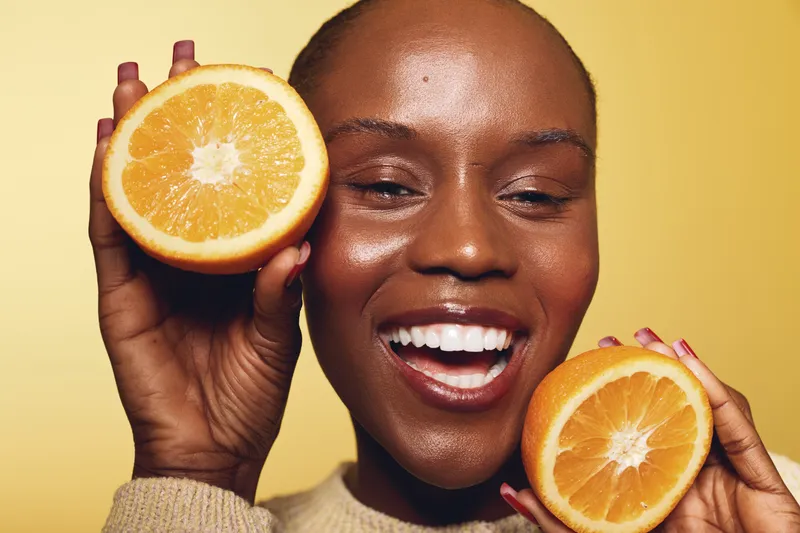 Happy African woman with dark skin holding two halves of fresh orange near her face, smiling with white teeth on yellow background. Concept of healthy lifestyle, fruit, and natural beauty.