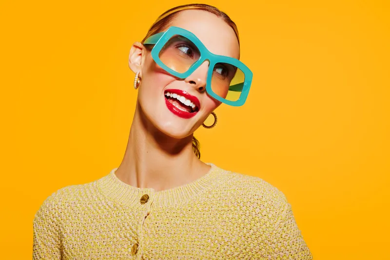Smiling woman with large blue sunglasses and red lipstick wears yellow knitted sweater. Bright orange background creates vibrant mood. Concept of fashion, style, confidence, and summer accessories.
