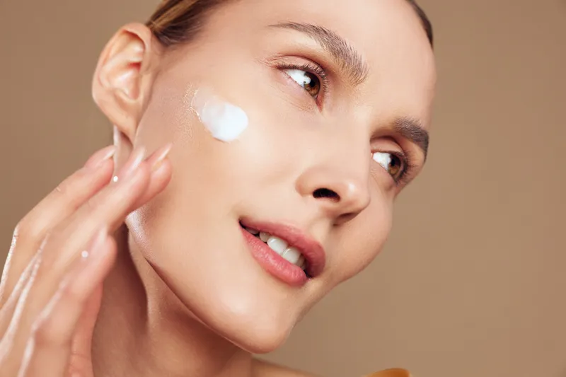 A young woman applies moisturizing cream on her clear skin with gentle fingers against a neutral background. Concept for skincare, beauty routine, and healthy complexion.