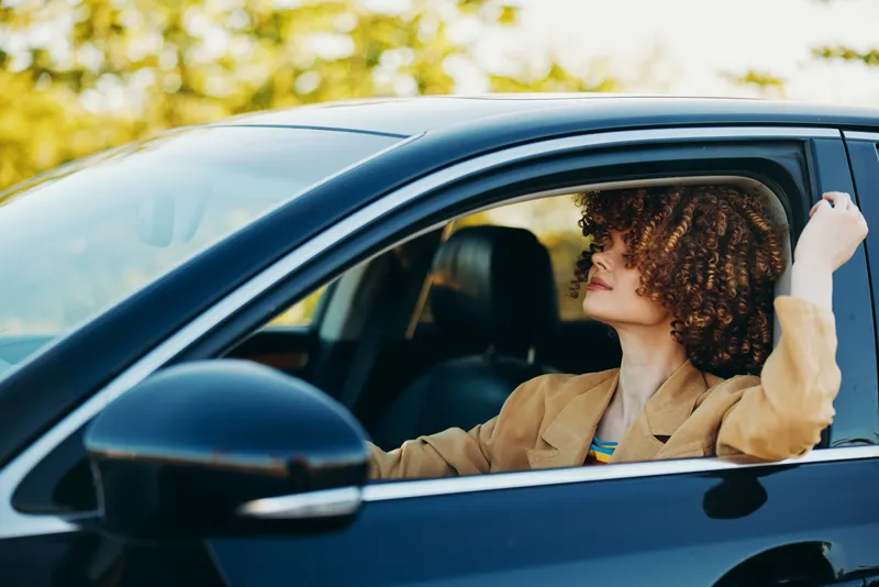 Young woman with curly hair sits behind the wheel of a dark sedan, glancing sideways with a relaxed smile as sunlit streets and green trees frame the moment