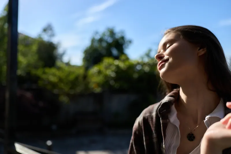 A smiling woman enjoys bright outdoor light, head tilted back, eyes closed, a casual lifestyle moment that conveys relaxation, confidence, and warmth in a sunny setting