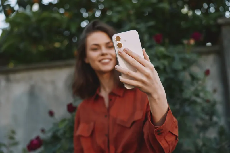 Woman lifestyle moment, taking a selfie with smartphone in a sunny garden, smiling, relaxed pose, candid portrait against green foliage and flowers