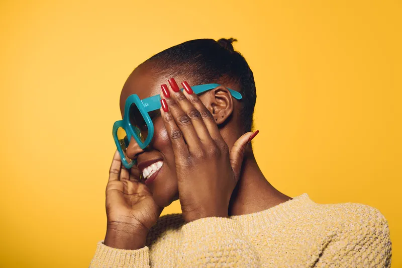 Happy African woman with dark skin adjusts blue sunglasses, smiling joyfully. She wears a beige sweater and has colorful manicured nails. Bright yellow background creates vibrant mood.