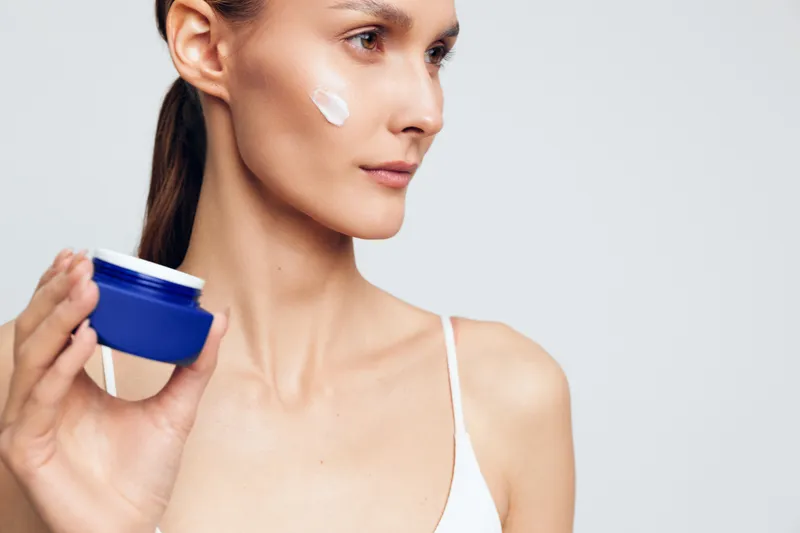 Woman skincare face cream moisturizer beauty wellness hygiene cosmetic care, woman applies white cream to cheek, holding blue jar, clean studio background, soft light