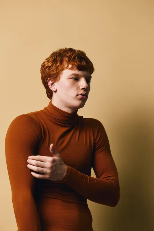 Young man with red hair wearing brown turtleneck sweater standing against beige background with thoughtful expression and hand crossed over chest in portrait style photo.
