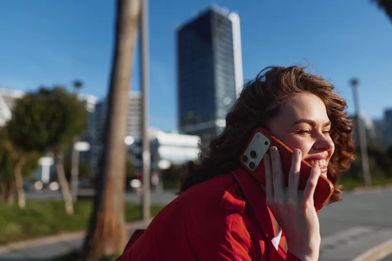 A young woman with curly brunette hair in a red jacket talks on a smartphone outdoors in an urban park on a sunny day, smiling as city buildings rise in the background.
