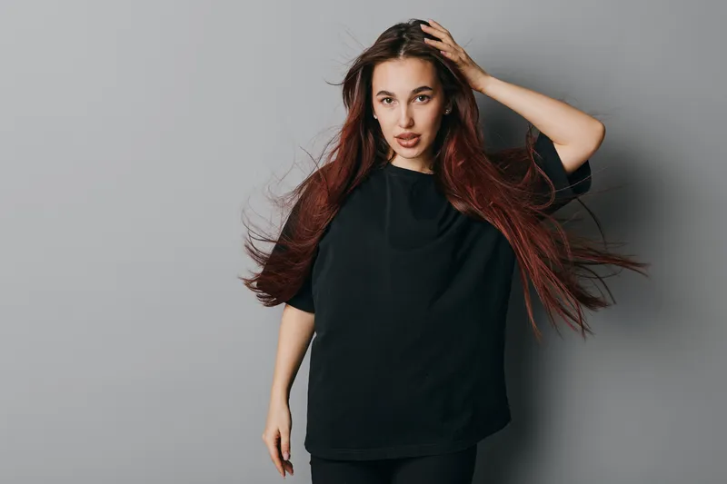 Young woman with long red hair and casual black t shirt posing with hand on head against gray background. Female model with confident expression and loose hairstyle in studio.