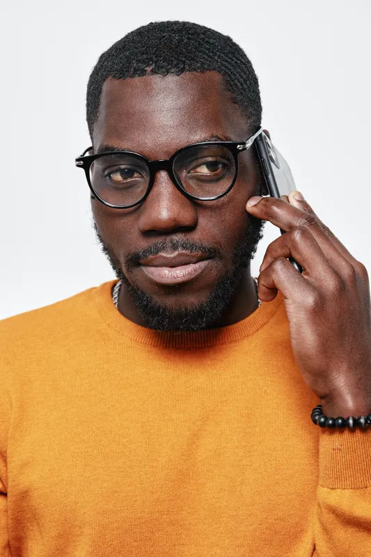 African American man with dark skin wearing glasses and orange sweater talking on smartphone, closeup portrait with serious expression on white background.
