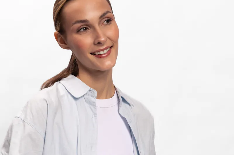 Young smiling woman with natural makeup looks away, wearing casual white shirt and t-shirt on white background. Concept of happiness, beauty, youth, and casual lifestyle.