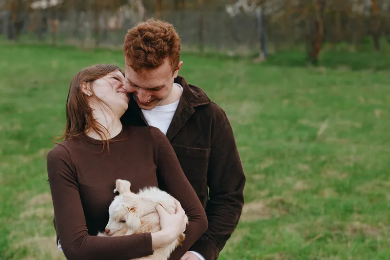 Couple smiling, cuddling a goat kid in a meadow. Casual clothing, affection and warmth define a relaxed lifestyle. Natural light highlights grass, sky, and a peaceful moment of love and togetherness.