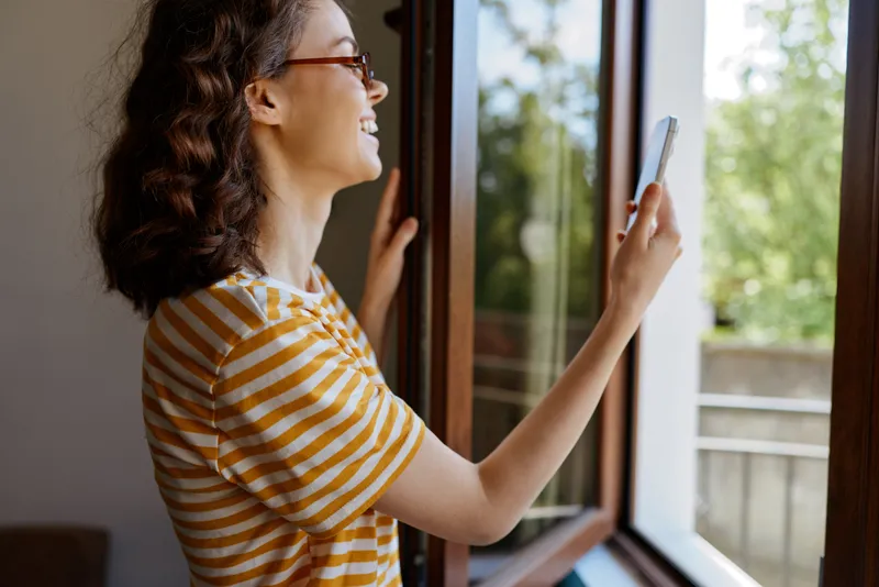 Young woman in a striped t shirt smiles broadly while holding a smartphone at an open window, enjoying a bright, cozy home moment.