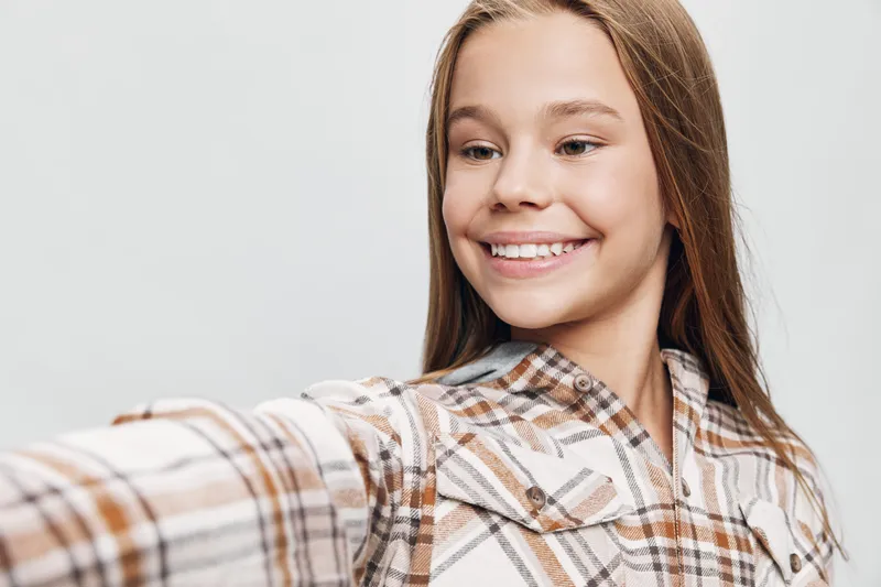 Teenage girl with long hair smiling and taking selfie on white background wearing plaid shirt with positive expression and natural makeup for social media content.