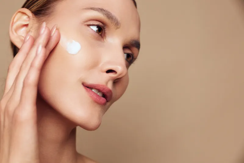 Woman applying skincare moisturizer to face in closeup beauty portrait, smooth skin, gentle hand touch, soft light, calm expression, beige clean studio background