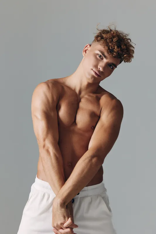 Young muscular man with curly hair posing shirtless in white shorts against gray background. Fitness, bodybuilder, toned physique and confident expression in studio lighting.
