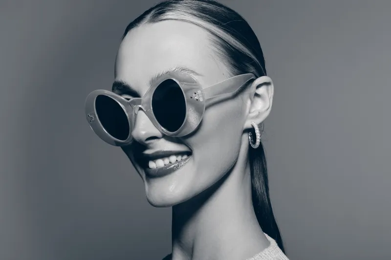 Smiling young Caucasian woman with fair skin wearing round sunglasses and hoop earrings, posing confidently against a plain light background.